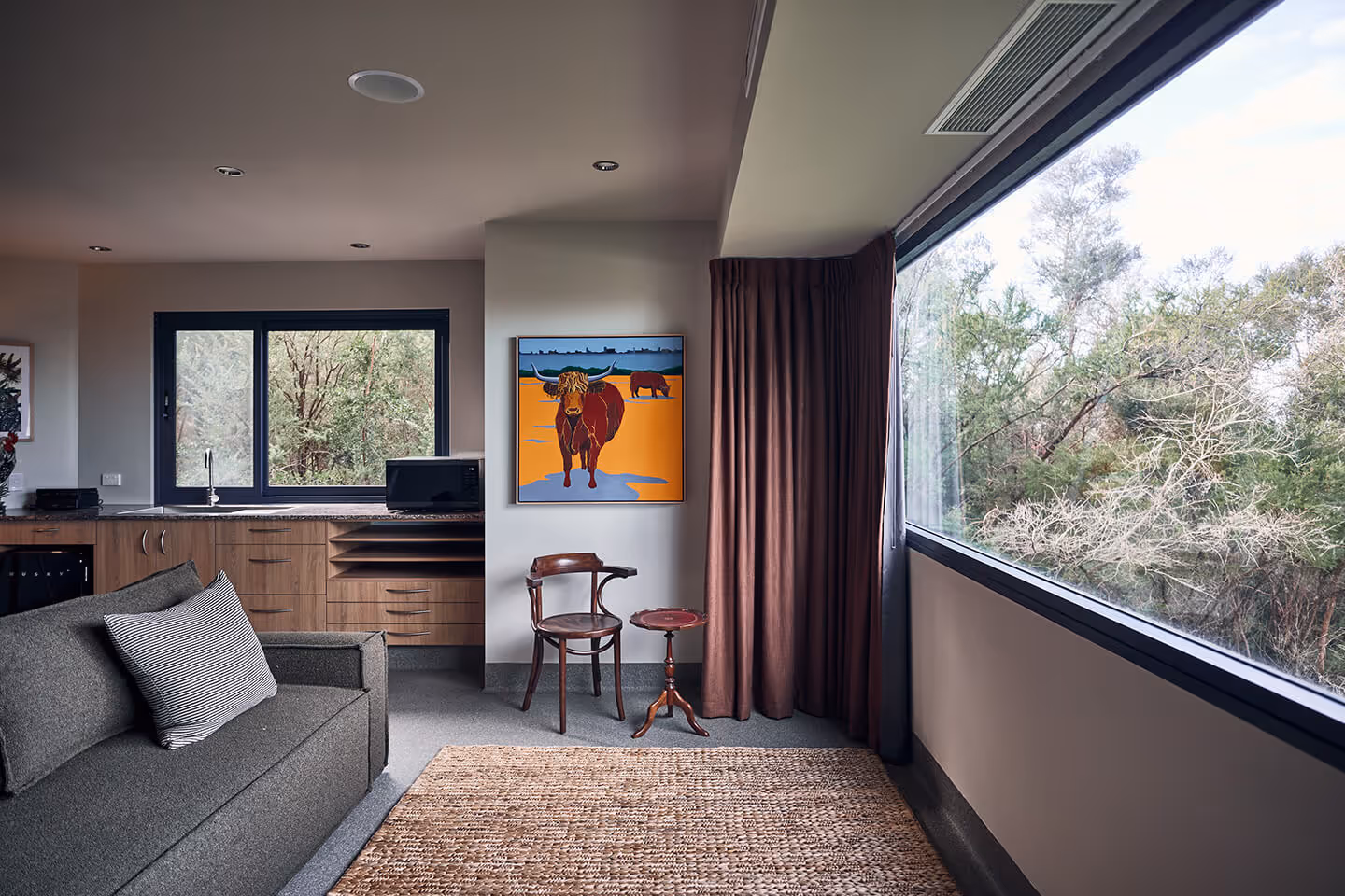 Modern living room with dark sofa, wooden chair, side table, large window, and colorful bull painting.