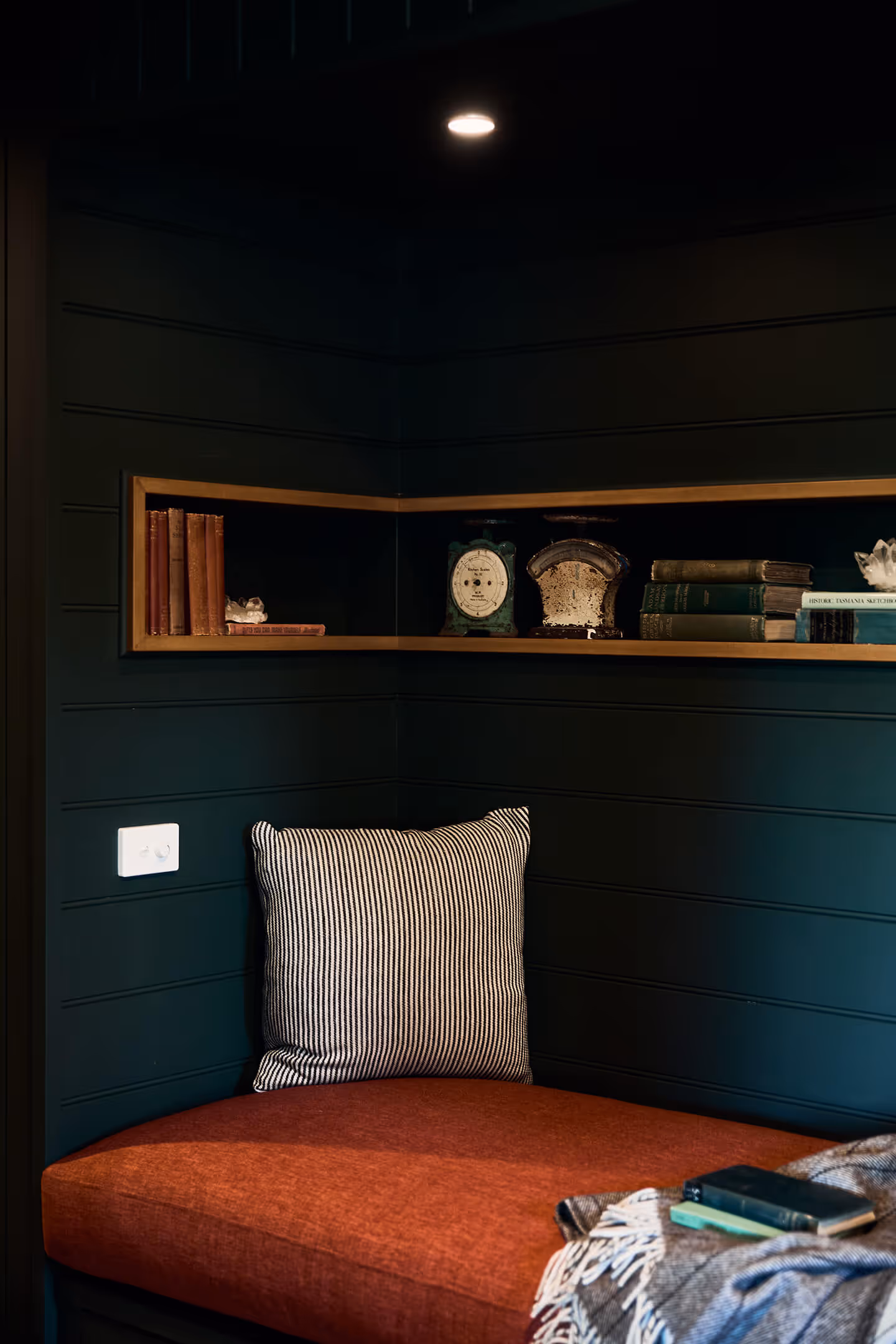 Cozy nook with orange cushion, striped pillow, blanket, and shelf with vintage books and scales.