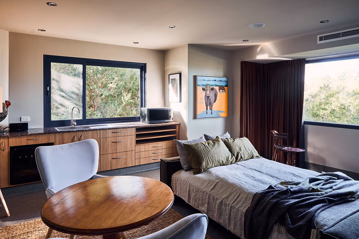 Cozy modern studio with bed, wooden table, chairs, kitchenette, large windows, and nature view outside.