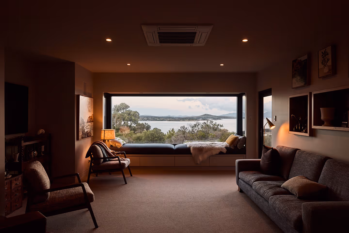 Cozy living room with a large window seat overlooking trees and water at dusk.