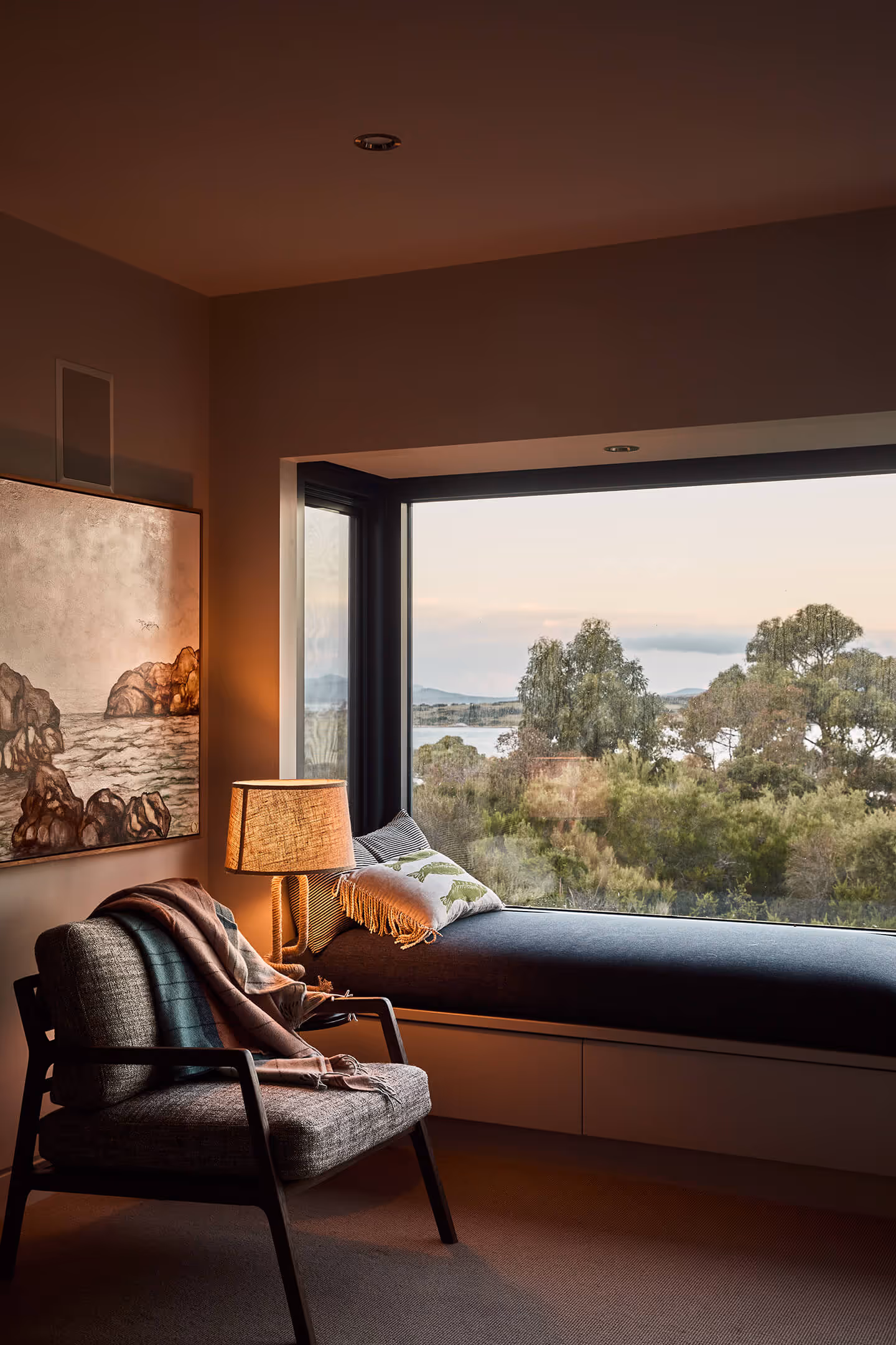 Cozy corner with armchair, throw blankets, lamp, and window seat overlooking trees and water view.