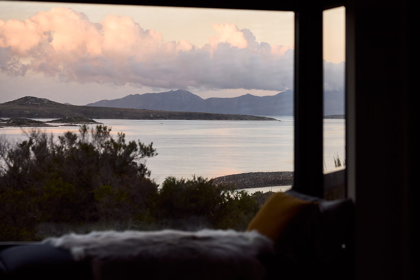 View of calm water, distant mountains, and clouds seen through a large window with a couch inside.