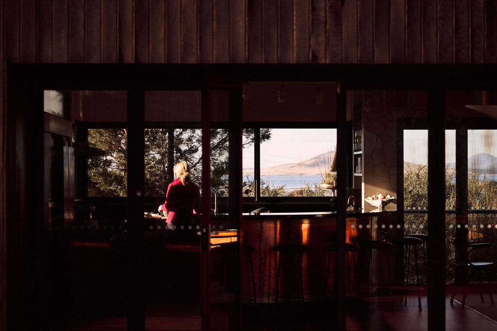 Woman in pink sweater standing in a wood-paneled kitchen with large windows showing trees and water view.