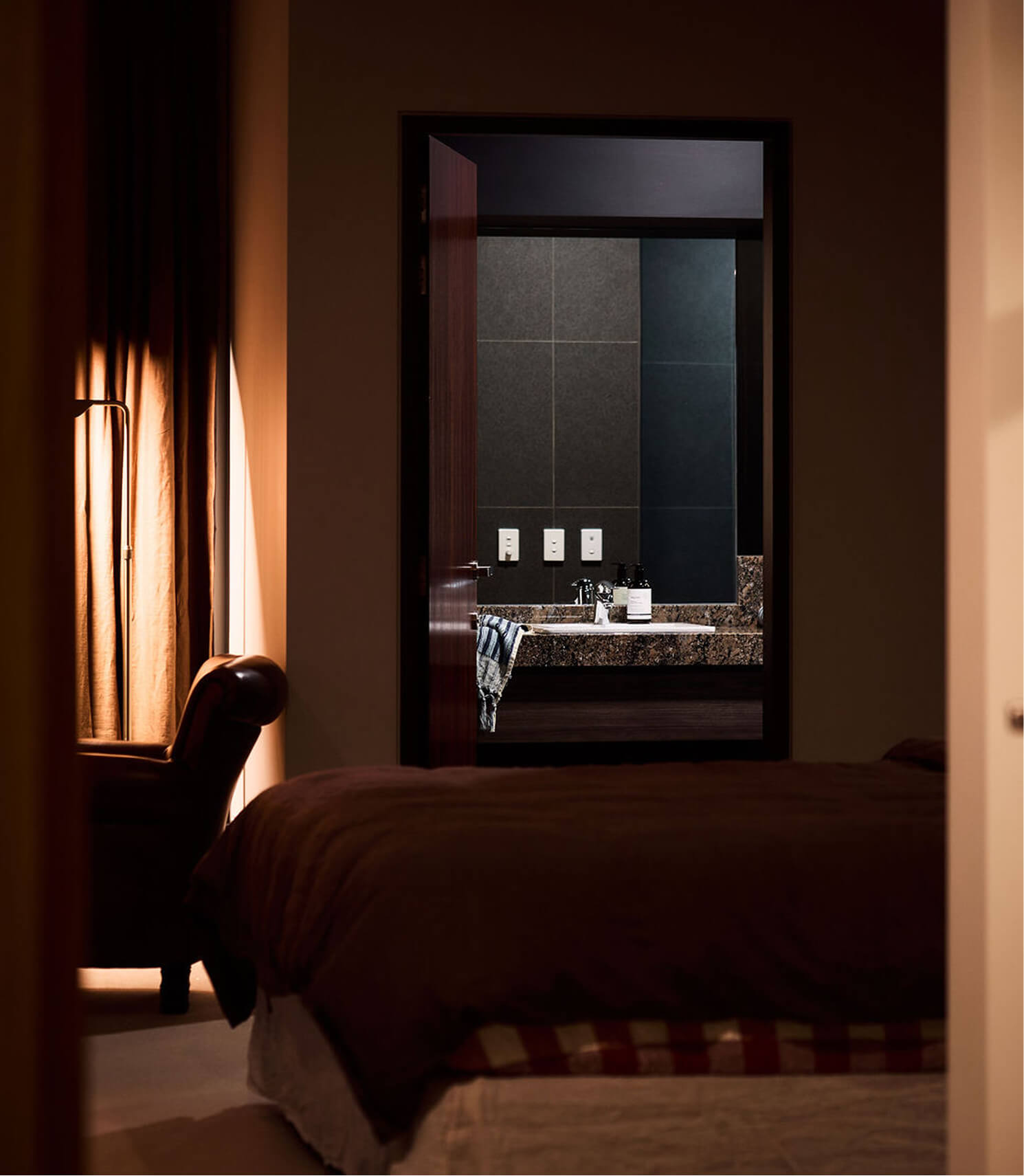 Dimly lit bedroom with bed and leather chair near window, looking into bathroom with sink and mirror.