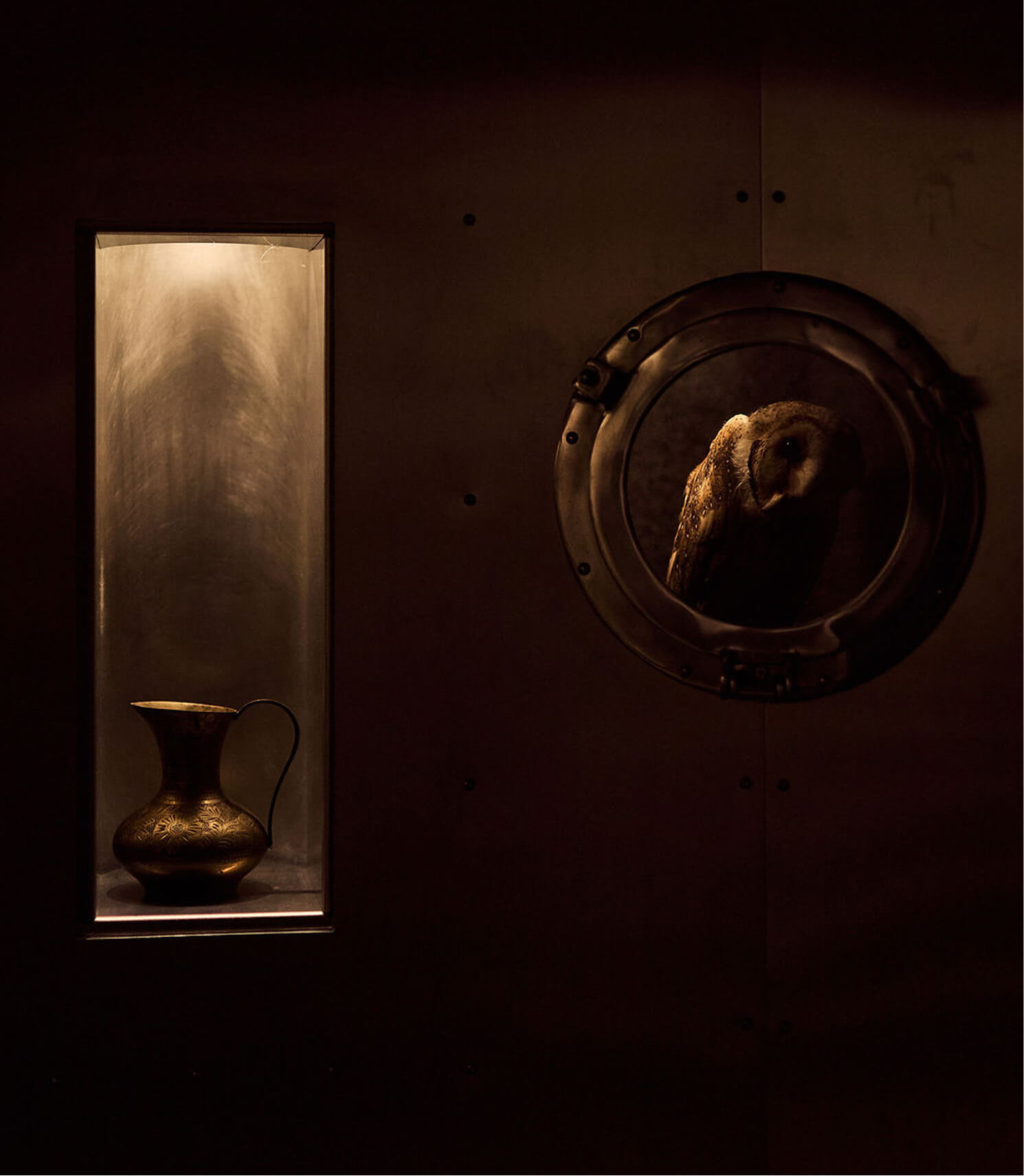 Dimly lit scene with an ornate bronze pitcher and an owl behind a circular porthole window.