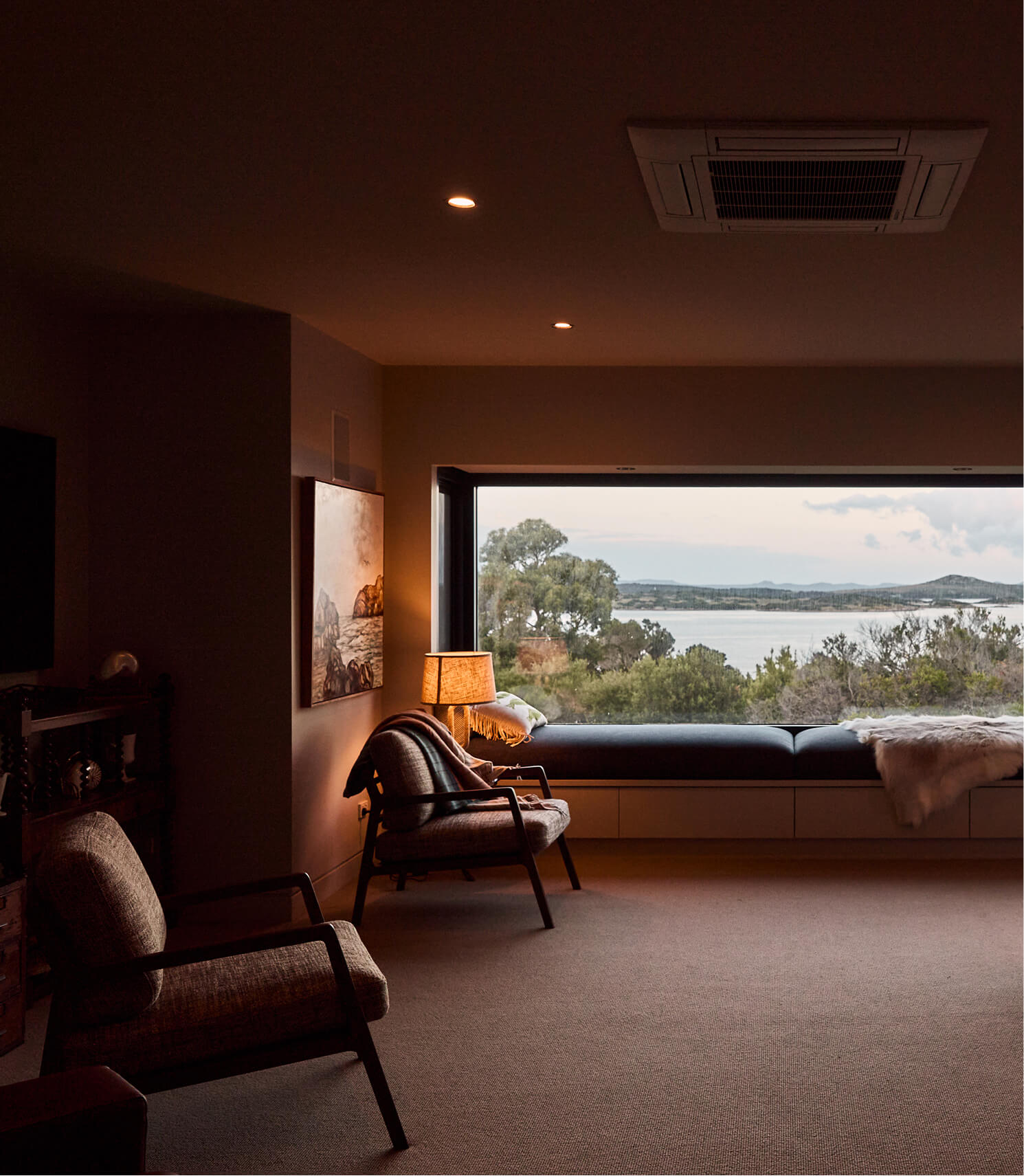 Cozy room with two chairs, lamp, and large window seat overlooking trees and water outside.