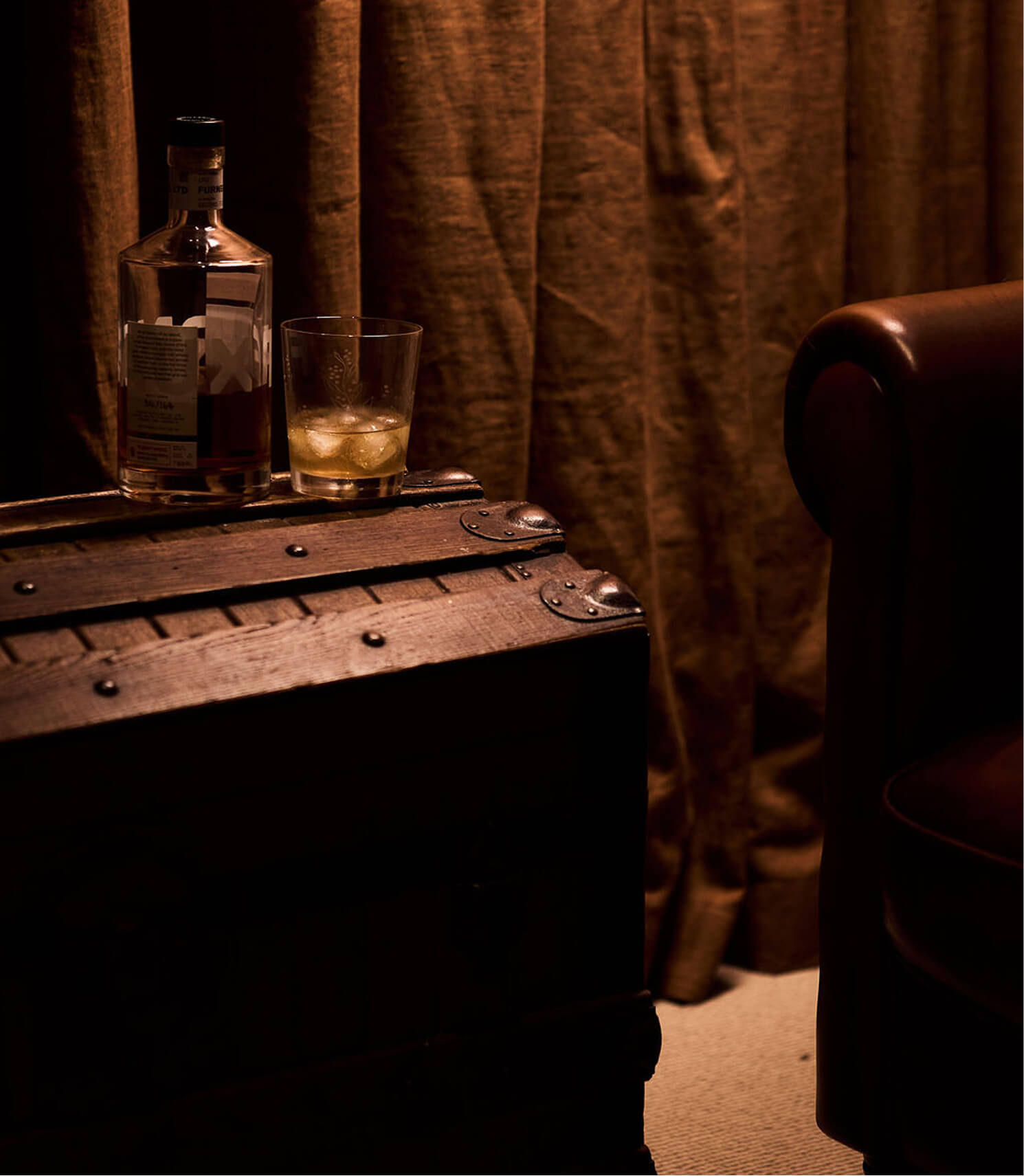 Glass of whiskey with ice and a partially filled bottle on a wooden trunk beside a brown leather chair.
