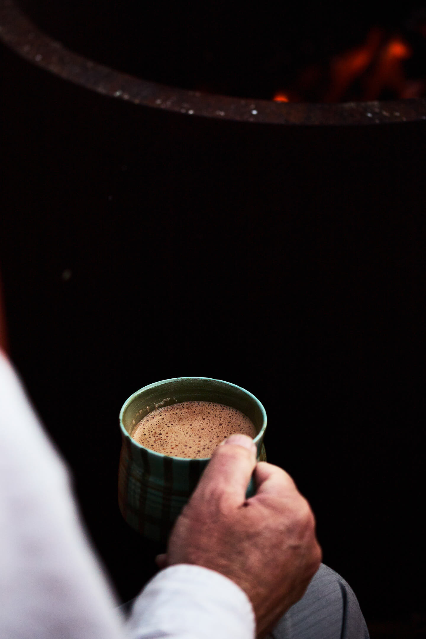 Close-up of a hand holding a green cup filled with frothy hot beverage in low light.