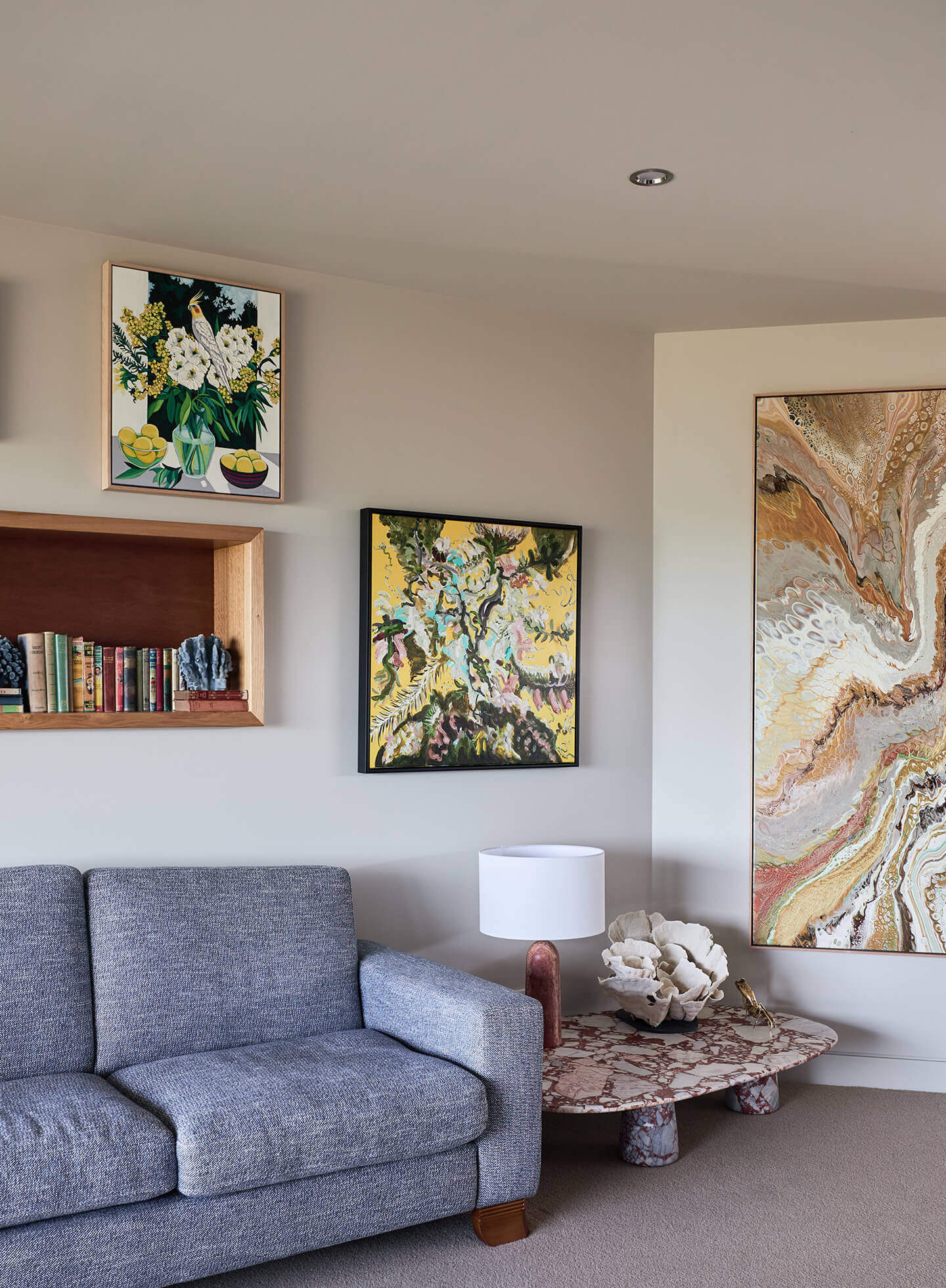 Living room corner with gray sofa, marble table lamp, abstract paintings, and a wooden shelf with books.