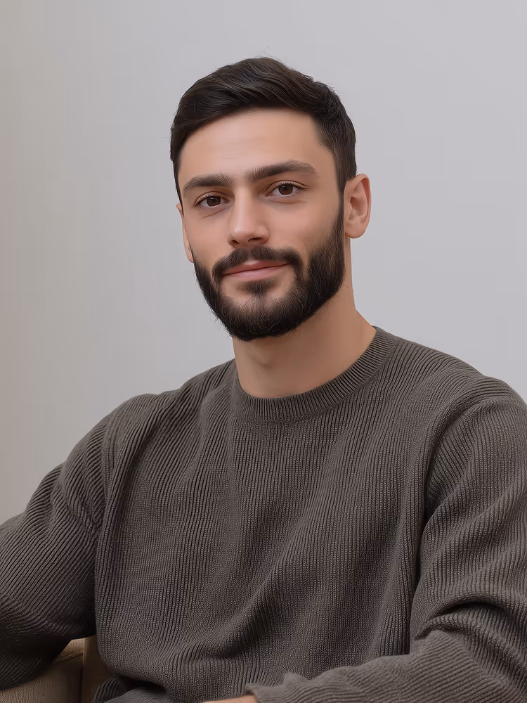 Man with dark hair and beard wearing a gray knitted sweater sitting and smiling against a light gray background.