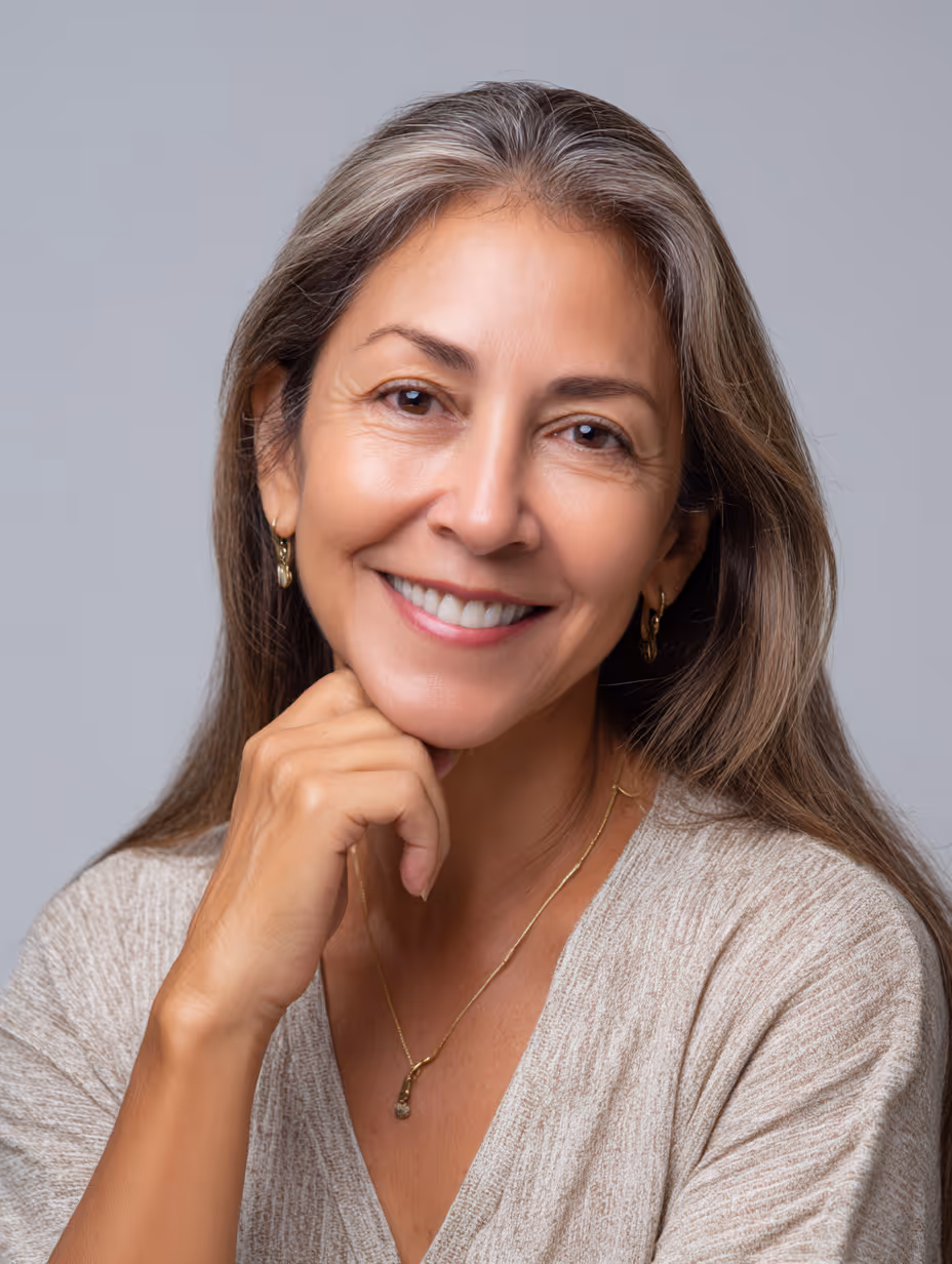 Smiling middle-aged woman with long gray hair resting her chin on her hand against a plain gray background.