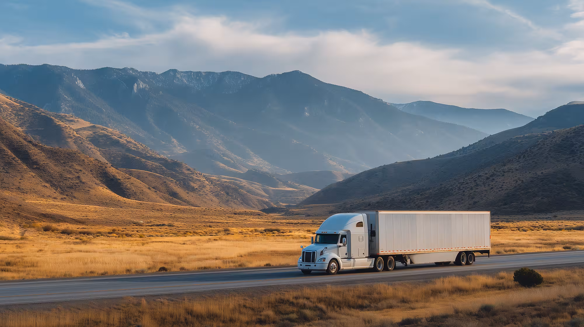 White semi-truck driving on a highway through a golden grassy valley with mountains in the background under a partly cloudy sky.