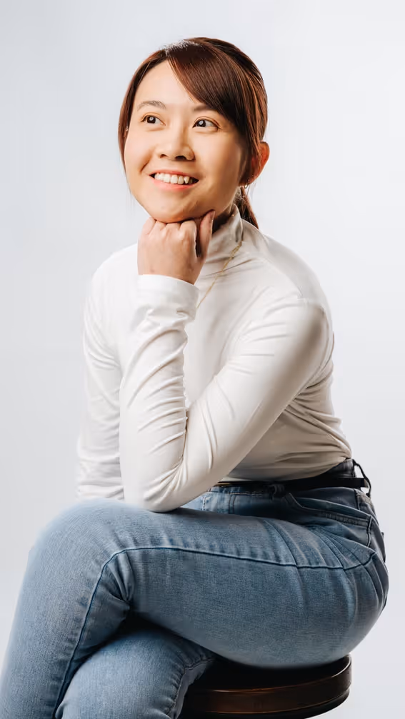 Smiling woman in white turtleneck and jeans sitting on a stool looking to the side.