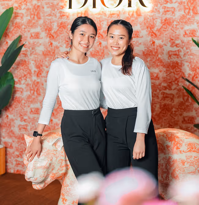 Two women smiling, wearing white tops and black pants, posing in front of a Dior sign and patterned wall.