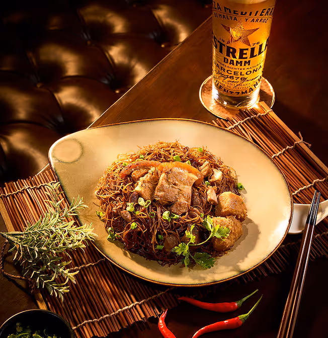Plate of stir-fried noodles with pork and herbs next to chopsticks and a glass of Estrella beer.