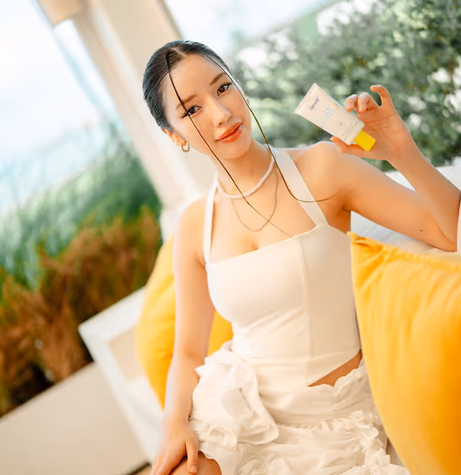 Woman in white dress sitting with yellow pillow, holding a small skincare product tube.