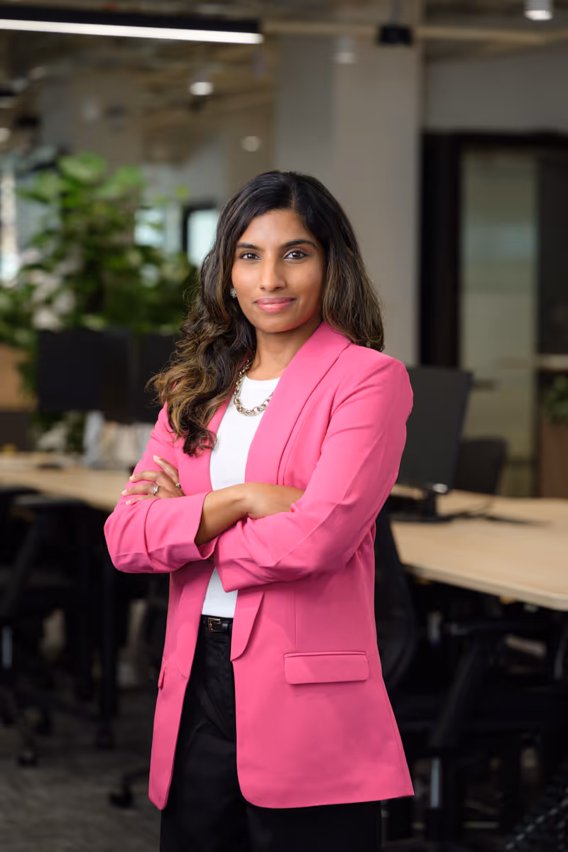 Confident woman with long hair wearing a pink blazer, standing with arms crossed in an office.