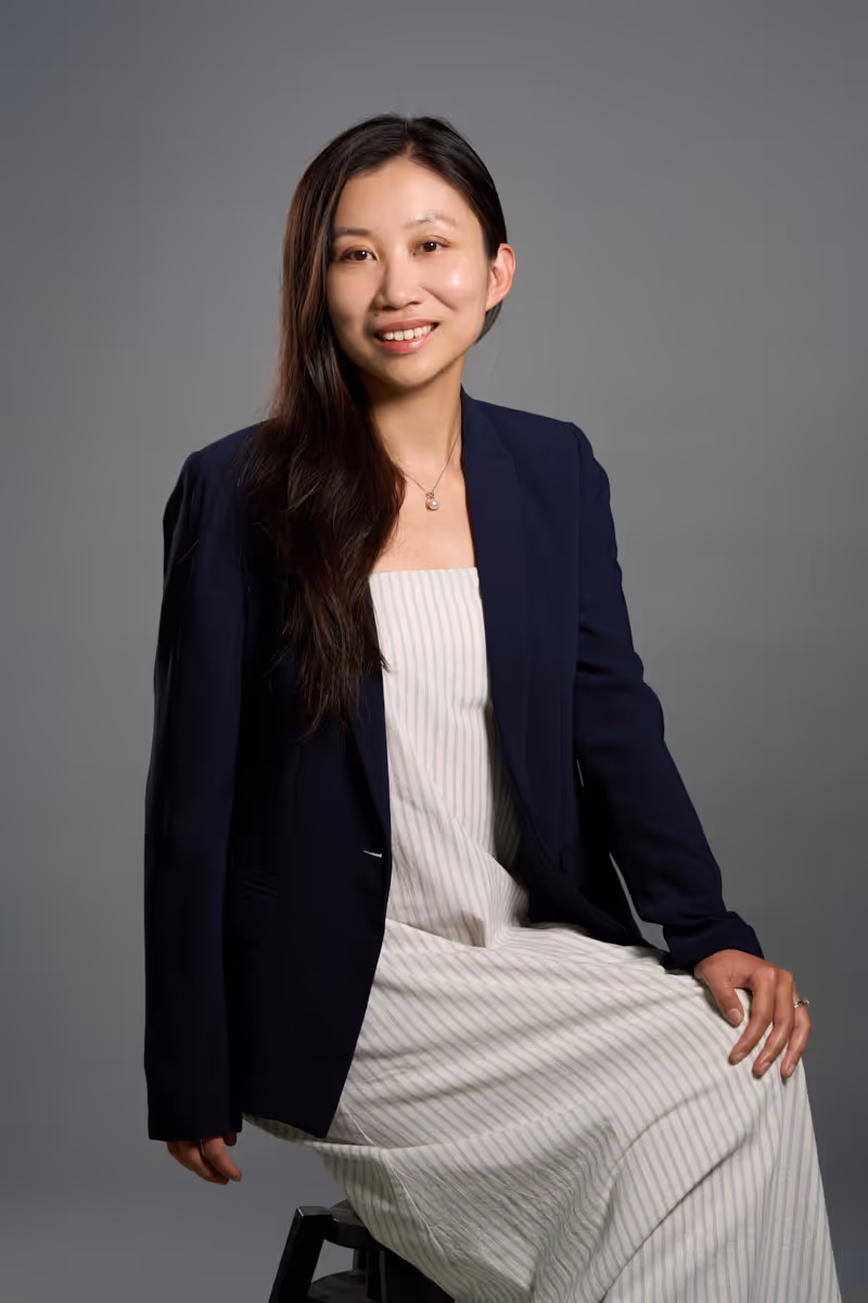 Smiling woman with long dark hair wearing a striped dress and navy blazer sitting on a stool.