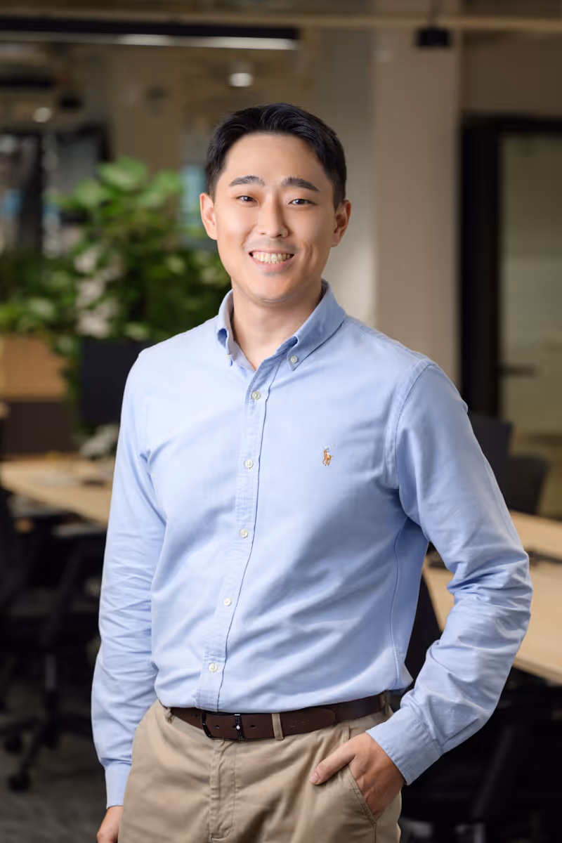 Smiling man in blue button-up shirt and beige pants standing in a modern office space.