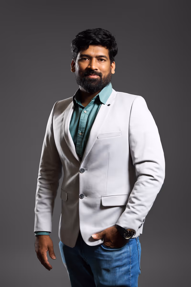 Bearded man in a white blazer and green shirt standing with one hand in pocket against gray background.