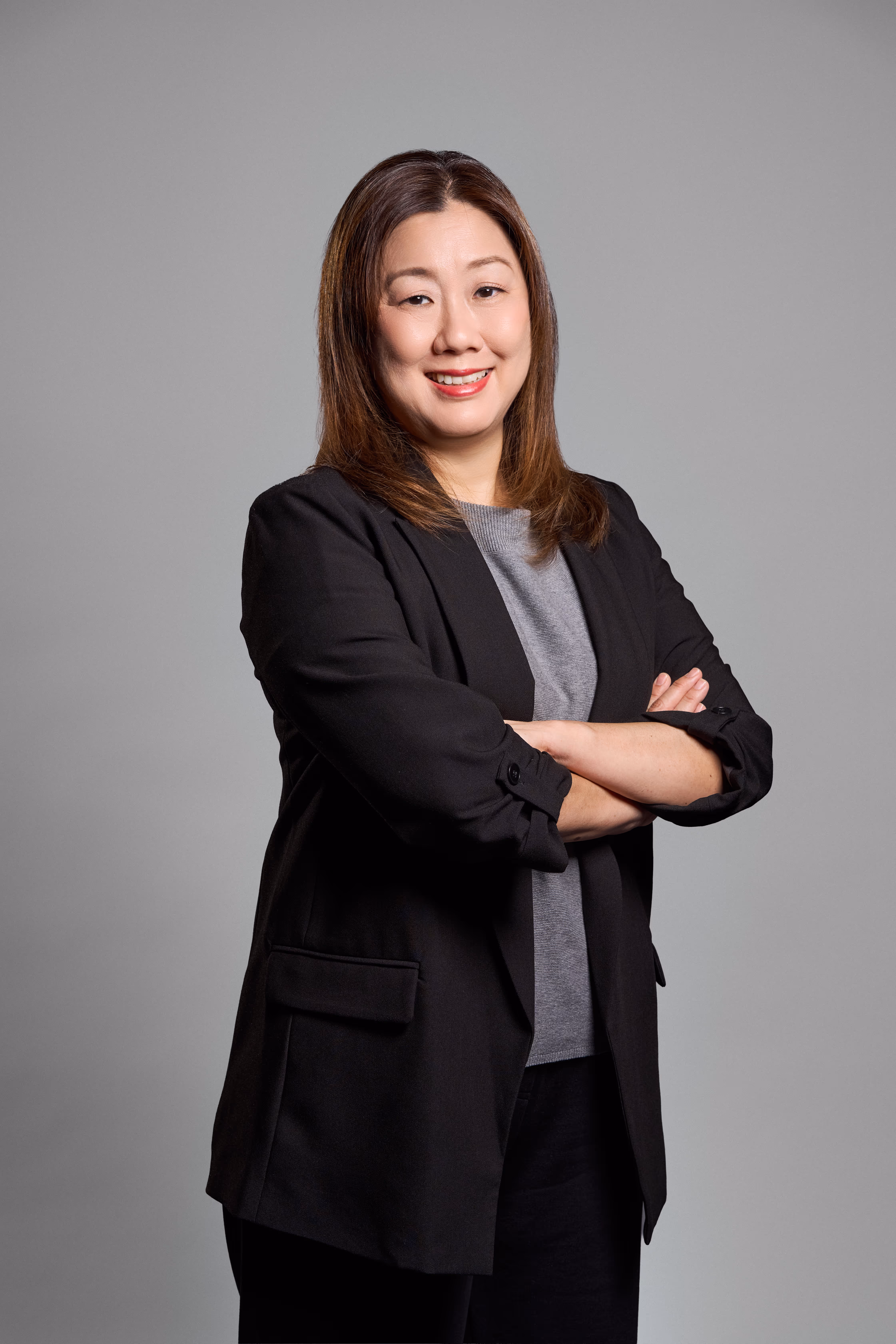 Smiling woman with straight brown hair in black blazer and gray top, arms crossed, gray background.
