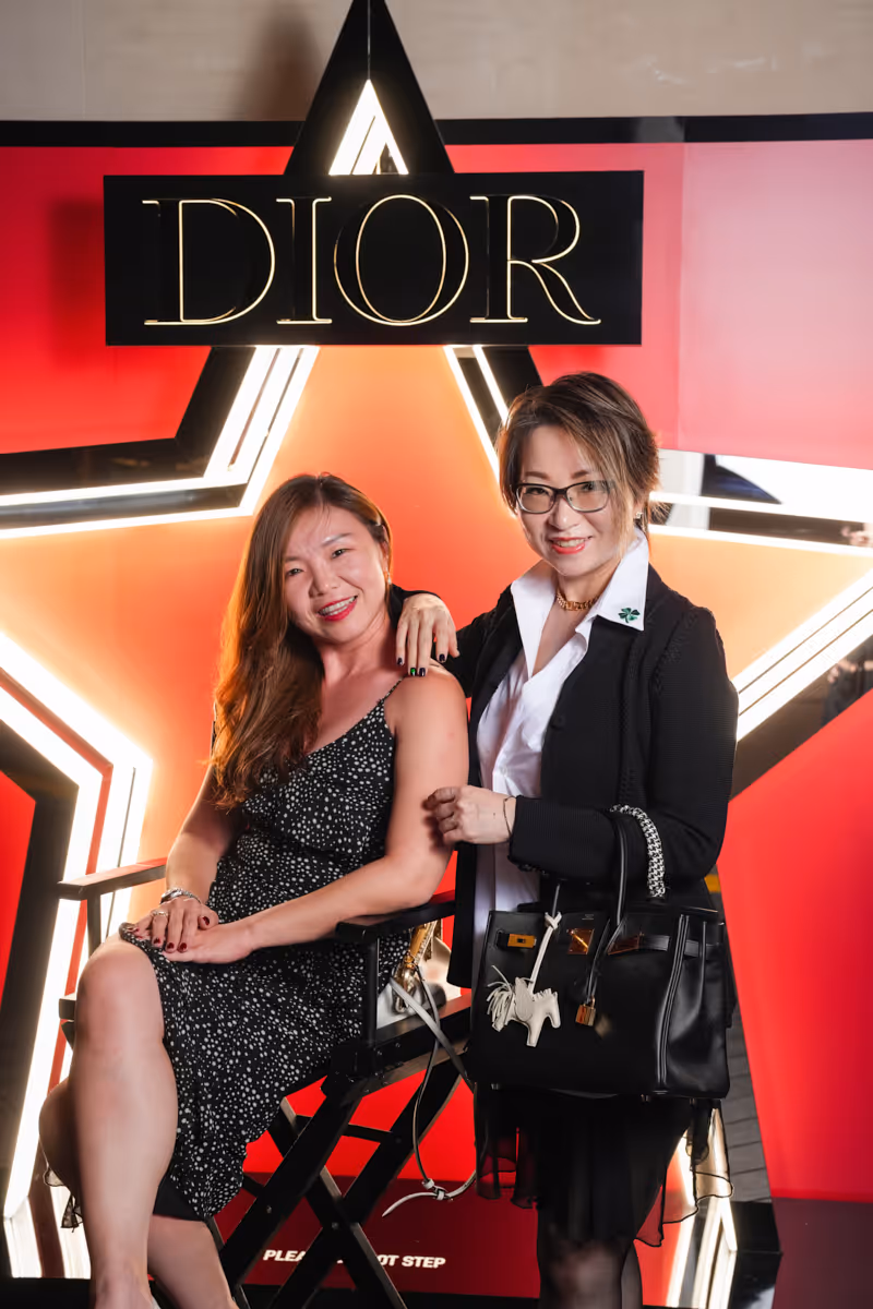 Two smiling women posing at Dior event with illuminated star and brand logo background.