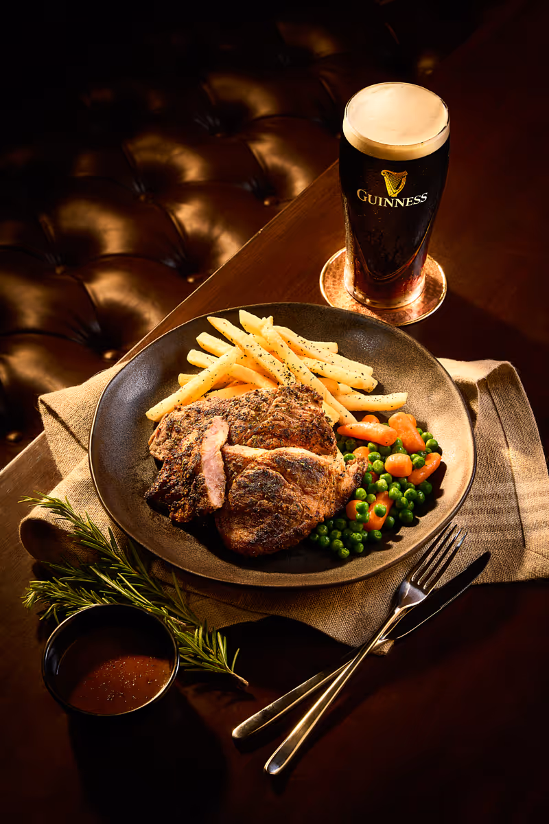 Plate with grilled meat, fries, peas, carrots, a cup of sauce, rosemary, and a pint of Guinness beer on a table.