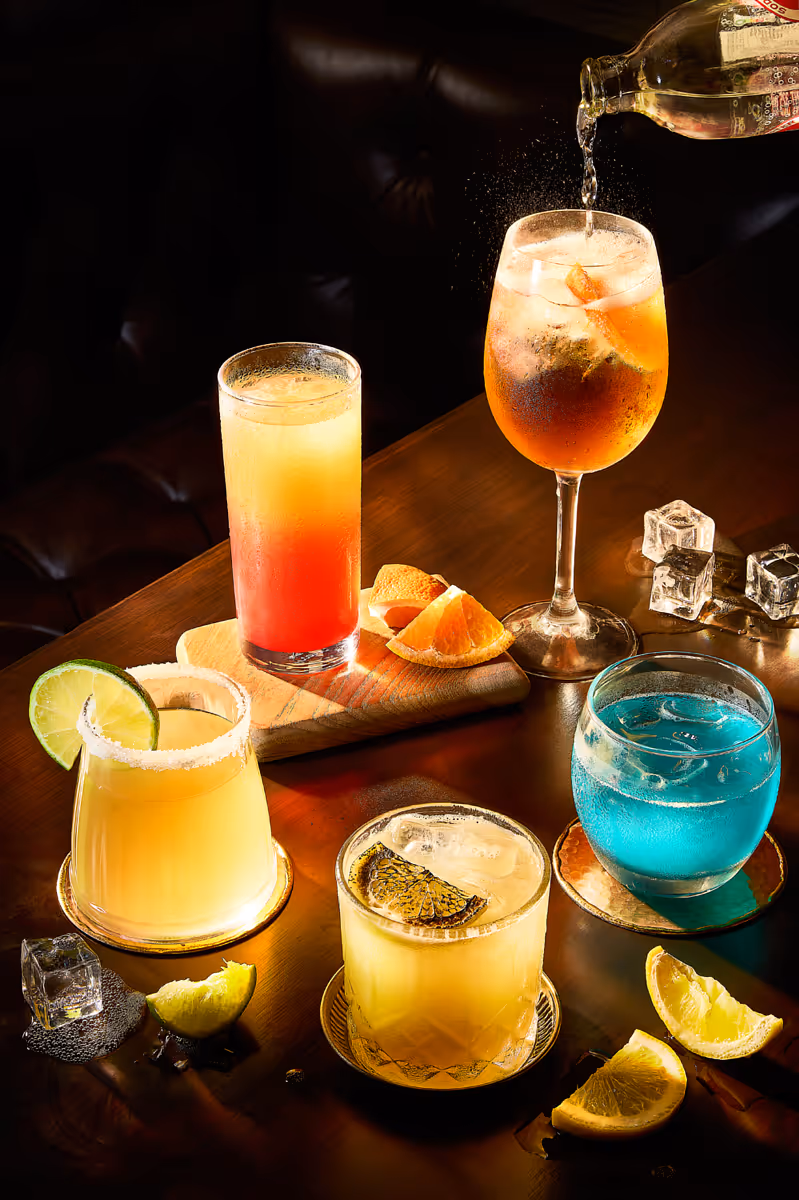 Variety of colorful cocktails on wooden table with citrus garnishes and ice cubes with soda pouring.