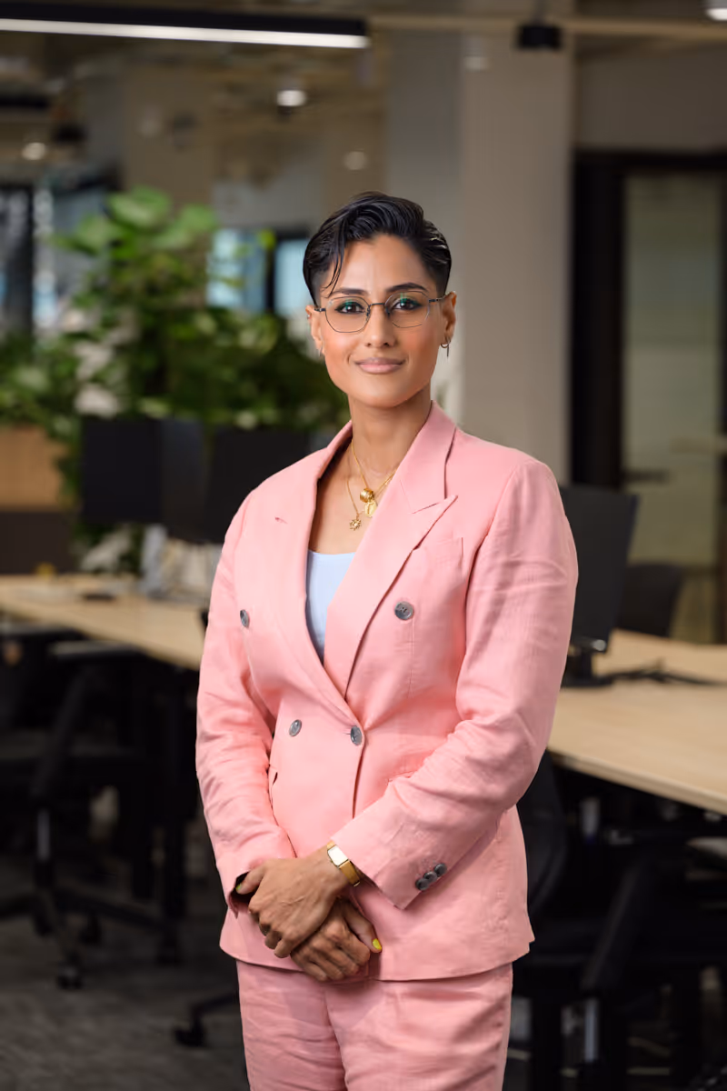Confident woman in glasses wearing a pink suit standing in an office with desks and plants.