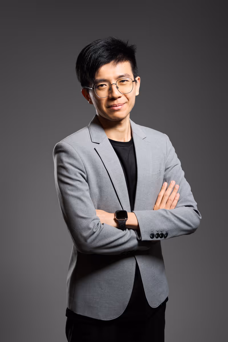 Man with glasses in a gray blazer and black shirt stands with arms crossed against a gray background.