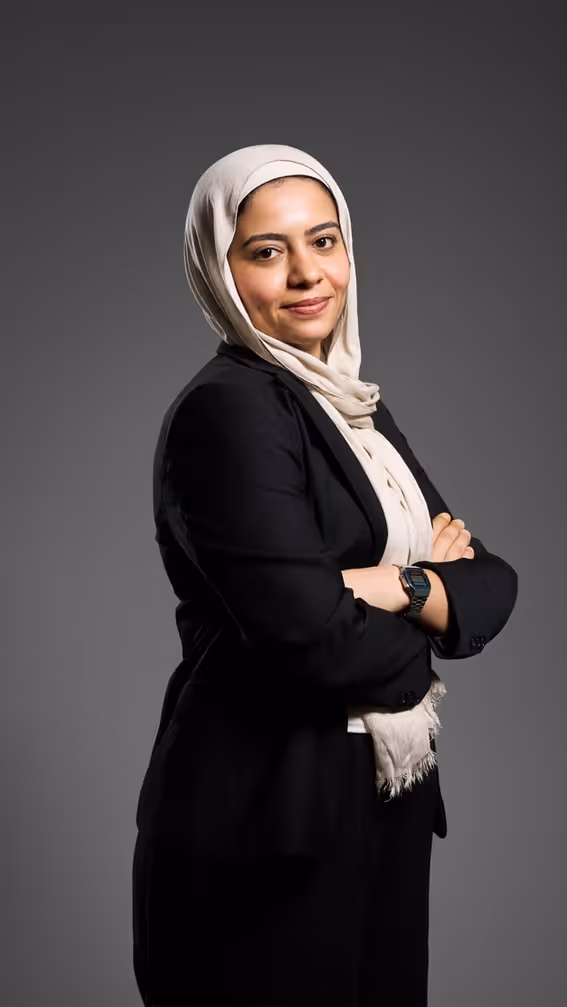 Confident woman wearing a light hijab and black blazer with arms crossed on a gray background.