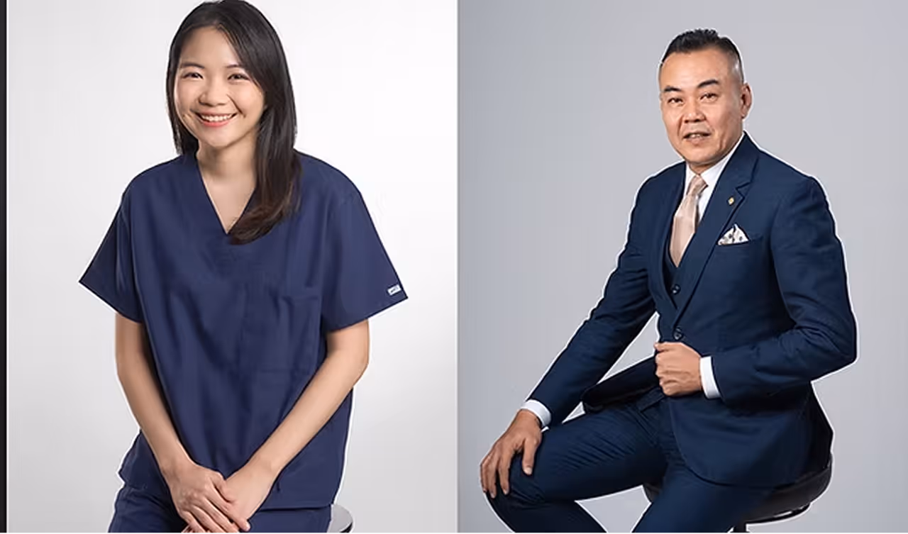 Smiling woman in navy scrubs and man in blue suit sitting on stools against a plain background.
