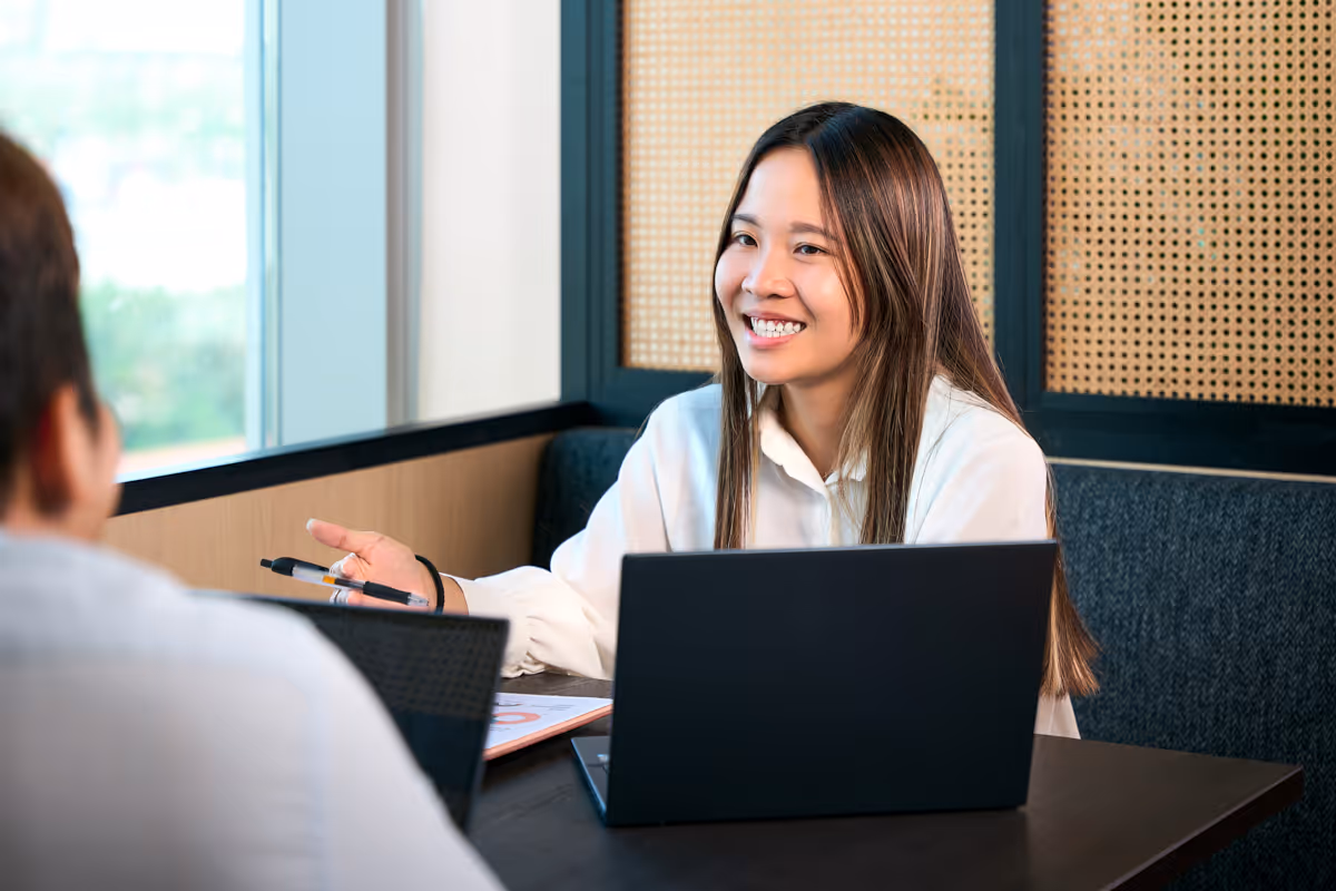 Woman in white shirt smiling and talking with a man across a table with laptops in an office.