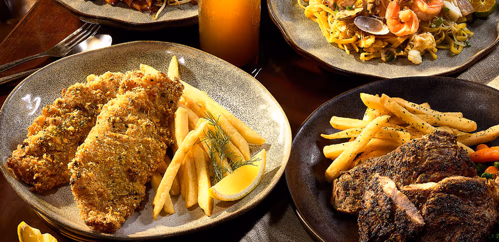Plates with fried fish, French fries, grilled steak, and seafood pasta with a glass of orange juice.