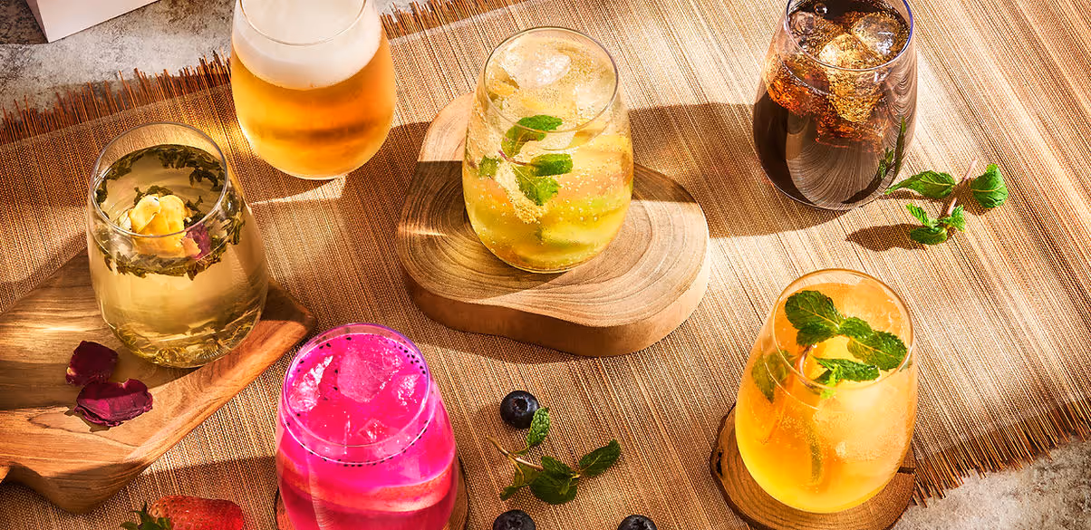 Six colorful drinks with ice and garnishes on wooden coasters and placemat with fruit and mint leaves.