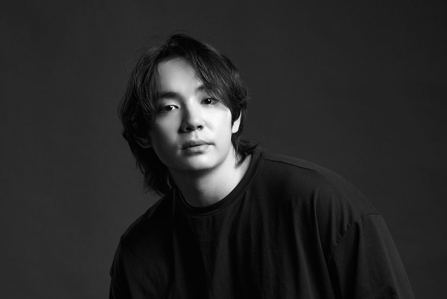 Black and white portrait of a young man with medium-length hair wearing a dark shirt against a dark background.