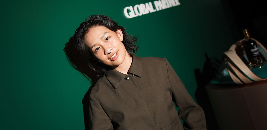 Smiling woman with short dark hair in brown shirt in front of a green wall with 'GLOBAL PARTNER' text.