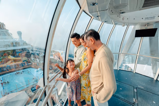 Four family members smiling and looking out large windows at a cruise ship pool deck.