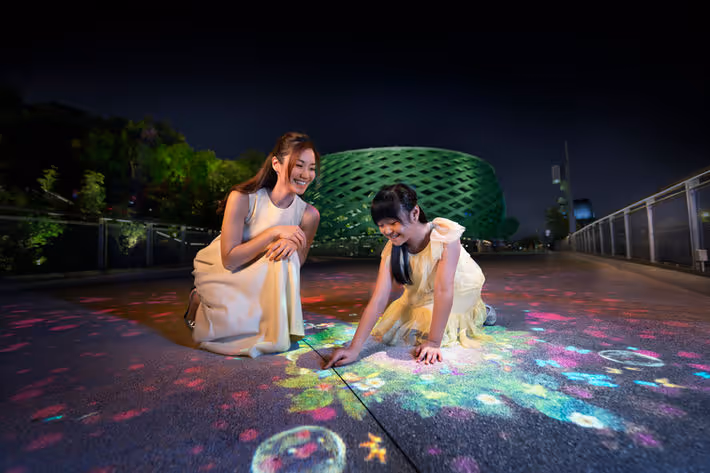 A woman and girl drawing glowing colorful patterns on the ground at night outdoors.