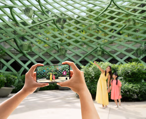 Hands holding smartphone capturing photo of woman in yellow and girl in pink under green lattice structure.