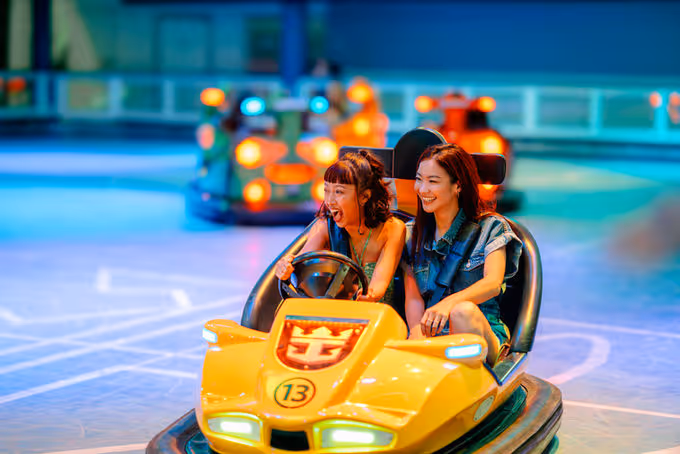 Two smiling women riding and laughing in a yellow bumper car at an indoor amusement park.