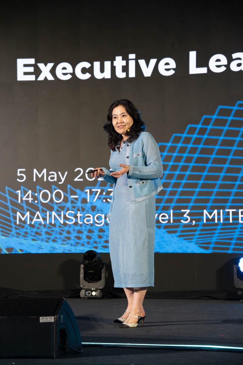 Woman in blue suit giving a presentation on stage with a screen showing event details behind her.