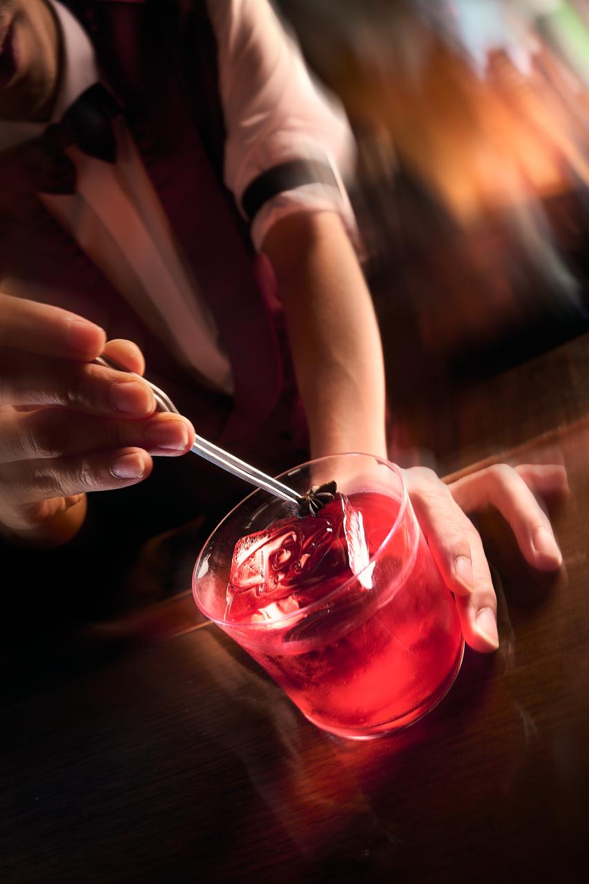 Person garnishing a red cocktail with a star anise using tweezers in dim lighting.