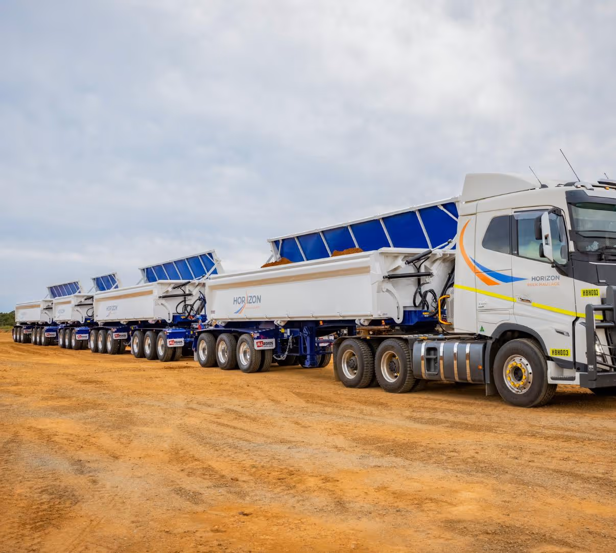 Line of connected white Horizon dump trucks with lifted blue dump beds on dirt ground.