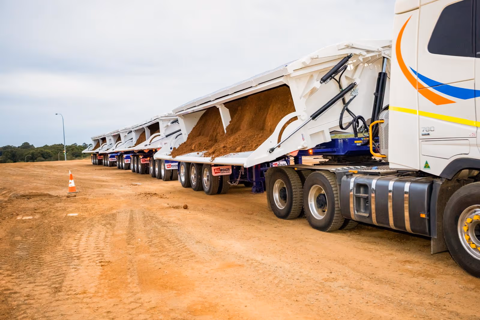 A fleet of white dump trucks unloading dirt on a construction site with a traffic cone nearby.