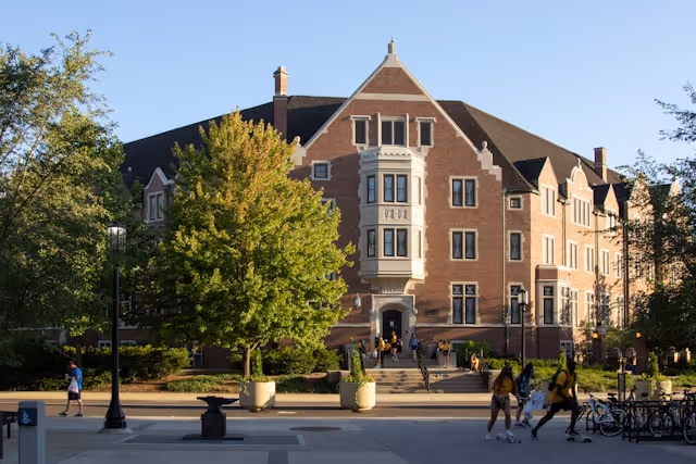 Brick academic building with bay windows, trees, and students walking on campus path at sunset.