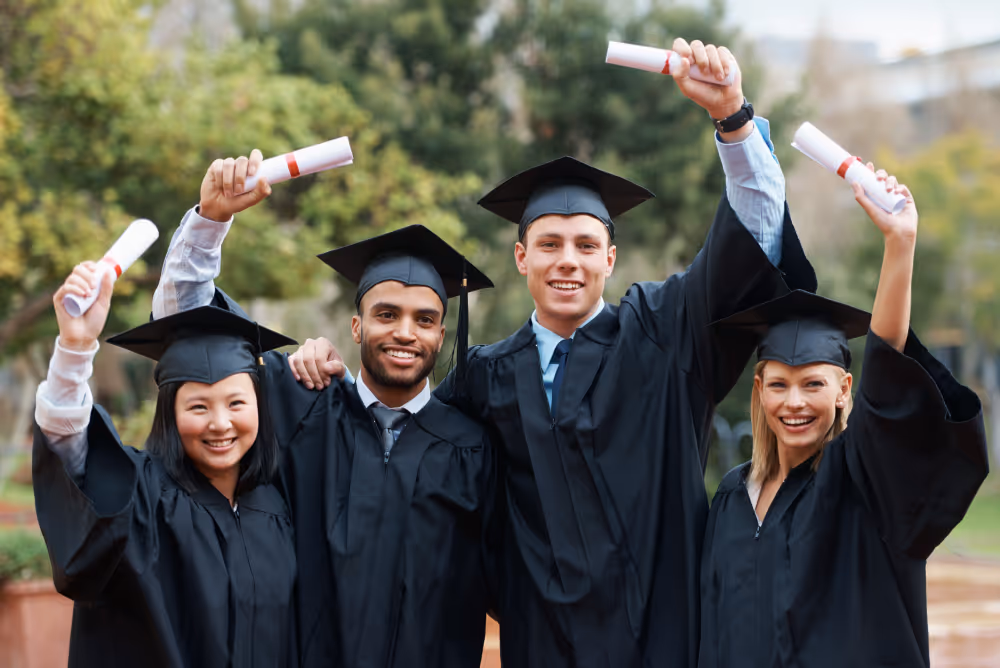Four diverse graduates in caps and gowns smiling and holding diplomas outdoors.