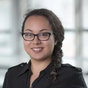 Smiling woman with glasses and a braid wearing a black top with a blurred background.