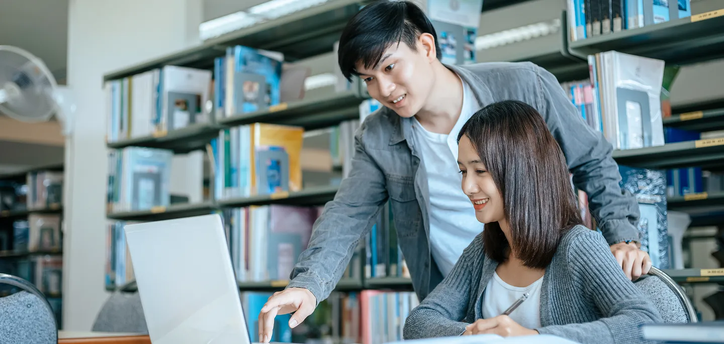 A man and woman studying together in a library, looking at a laptop screen with bookshelves behind them.