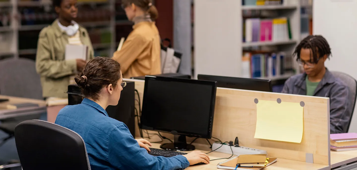 People working and talking in a modern library with computers and books on desks and shelves.